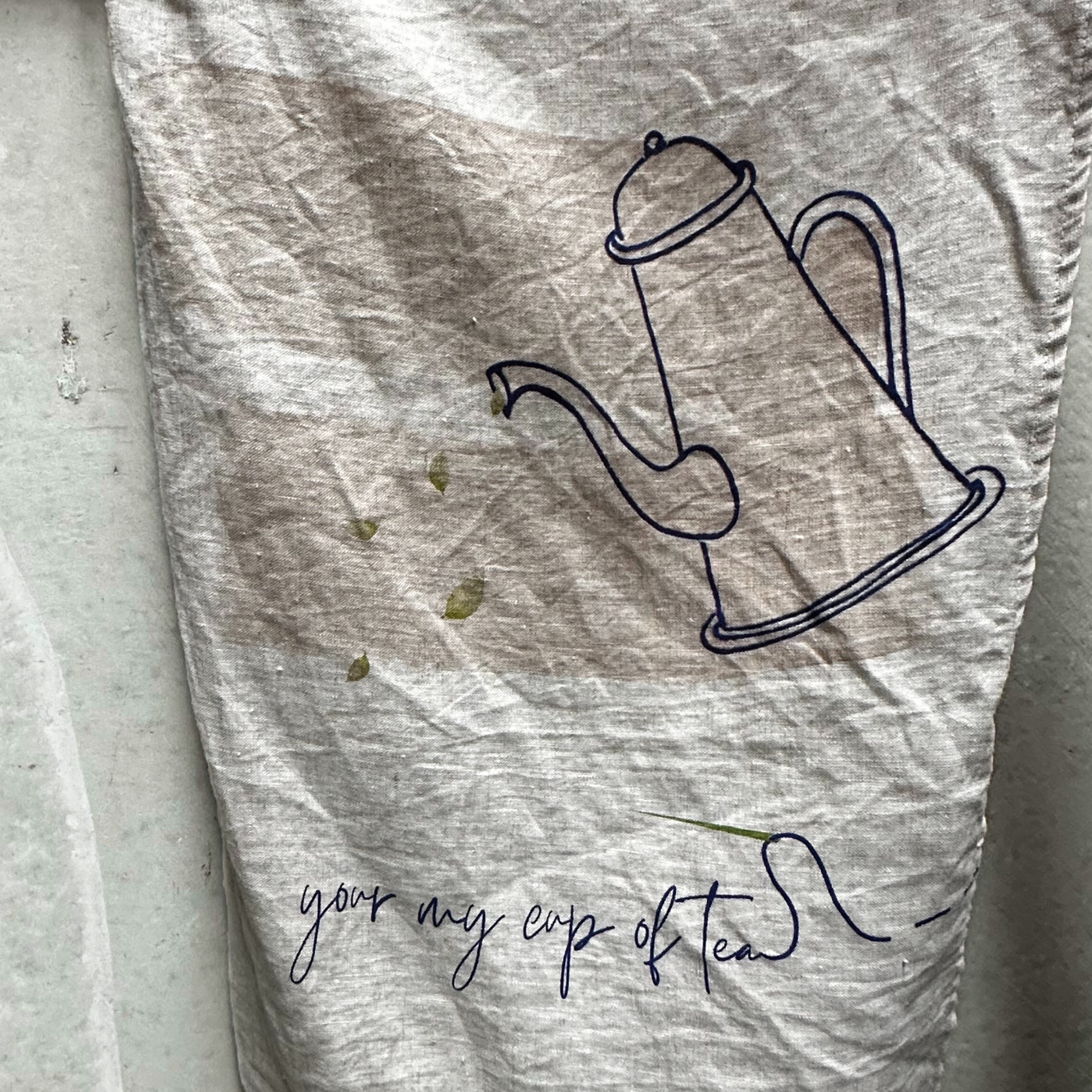 French linen tea towel with a teapot design and text 'your my cup of tea' on a textured surface.