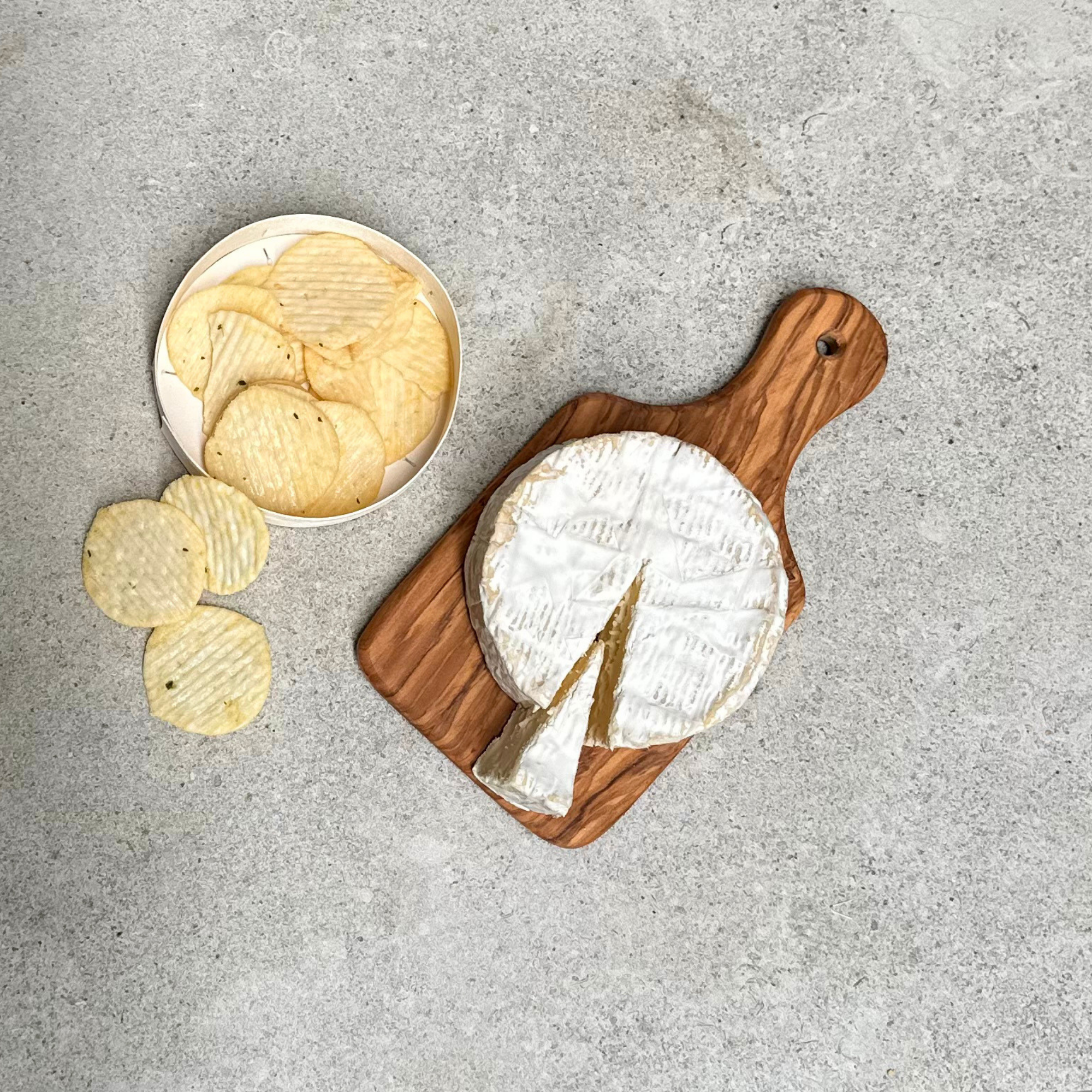 Solid olivewood chopping and serving board with handle, handcrafted in Germany, styled with Brie cheese and potato crisps.